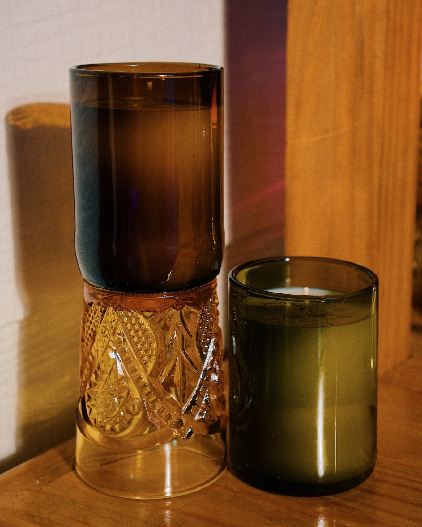 Two repurposed glass candle jars on a wooden surface, one antique green and one amber, set against a warm wood and white wall background.