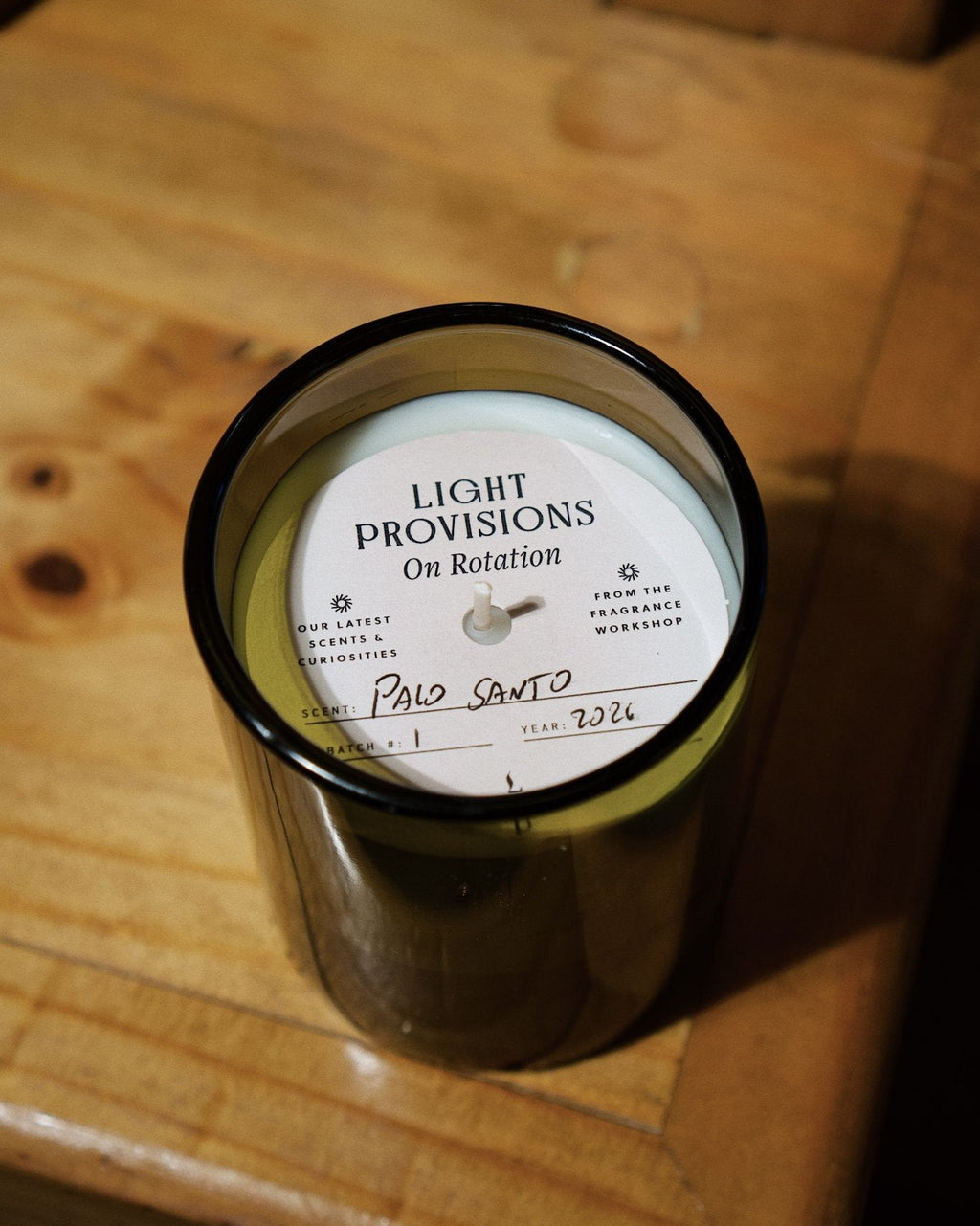 A dark amber glass jar candle with a white circular label reading 'LIGHT PROVISIONS On Rotation' and 'Palo Santo' written in script, placed on a wooden surface.