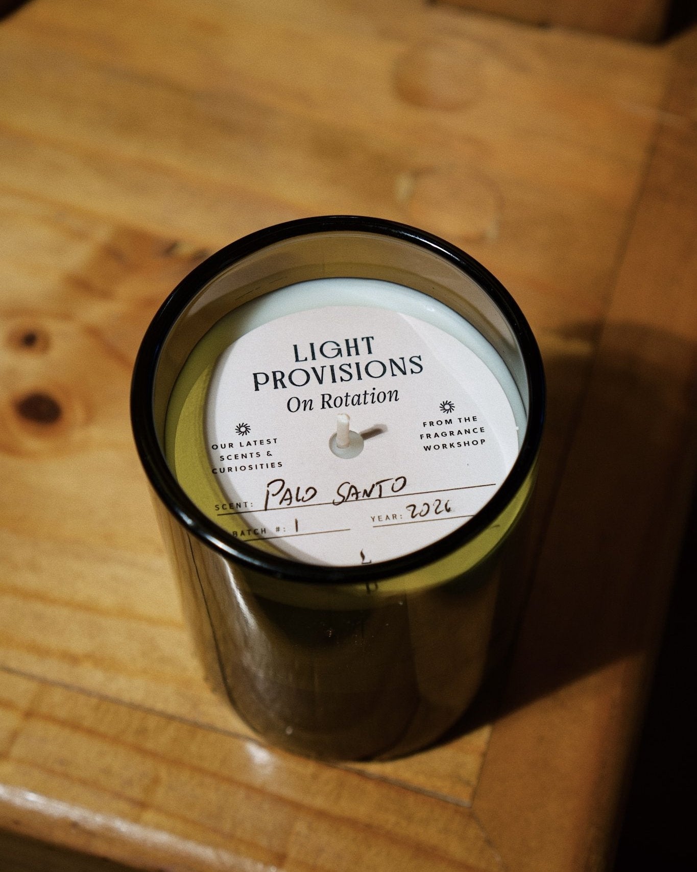 A dark amber glass jar candle with a white circular label reading 'LIGHT PROVISIONS On Rotation' and 'Palo Santo' written in script, placed on a wooden surface.