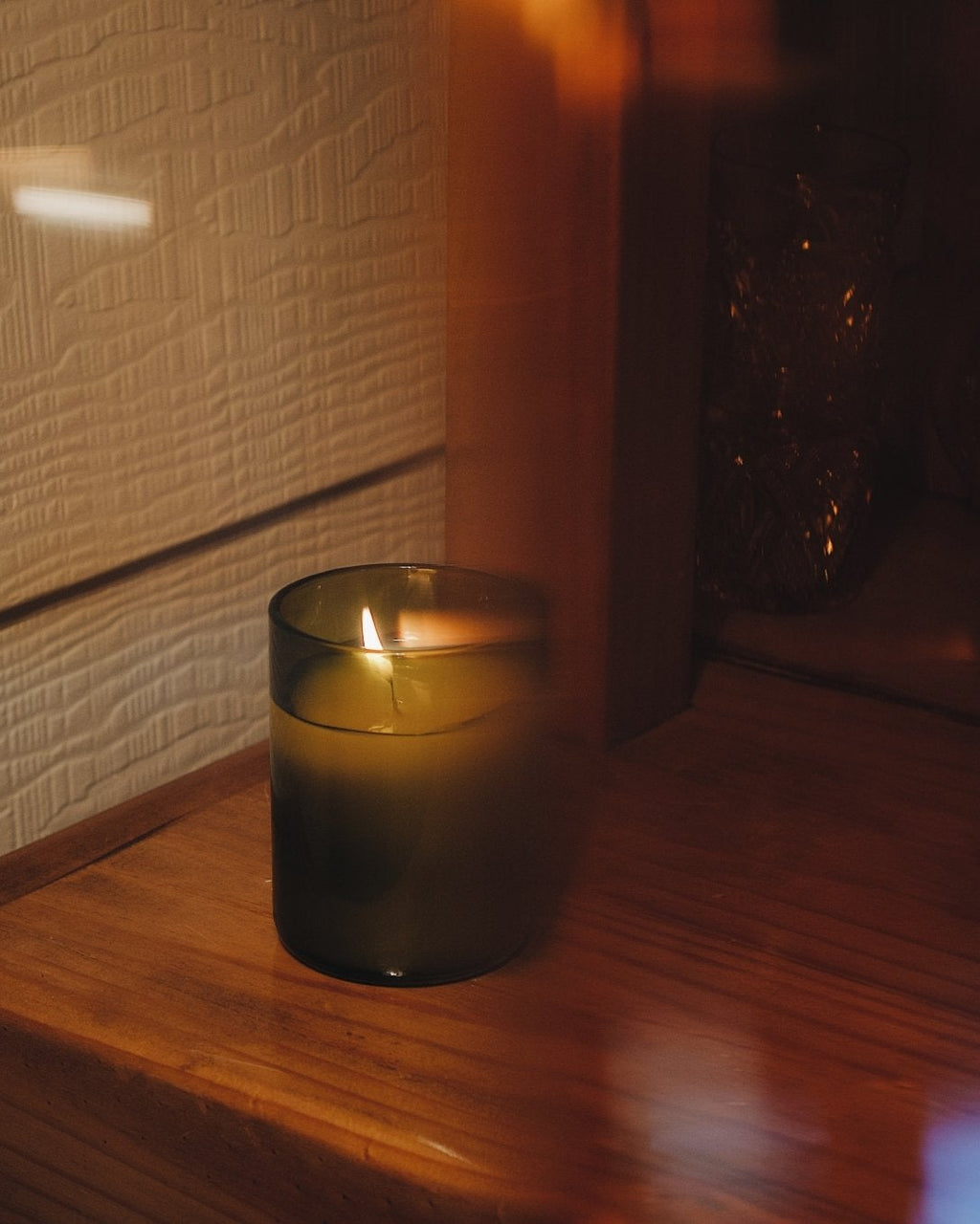 A lit candle in a dark amber recycled glass jar sits on a wooden surface next to a textured cream wall, captured in warm, moody lighting.