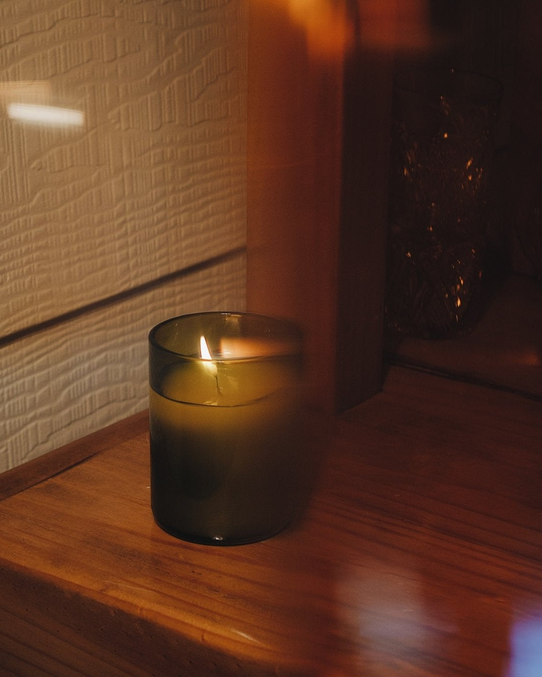 A lit candle in a dark amber recycled glass jar sits on a wooden surface next to a textured cream wall, captured in warm, moody lighting.