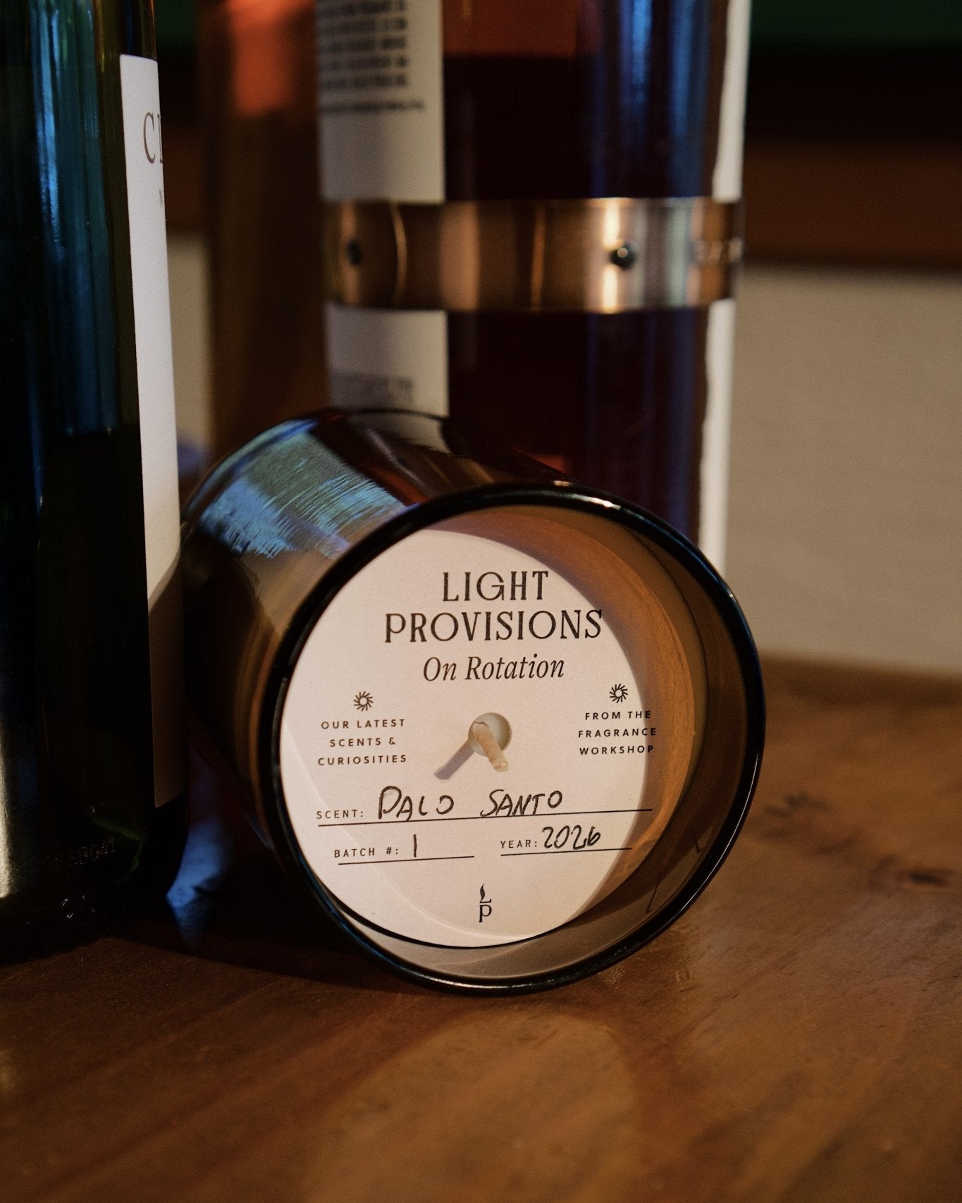 A dark amber glass jar candle lying on its side on a wooden surface, showing the circular white label that reads 'LIGHT PROVISIONS On Rotation' and 'Palo Santo'.
