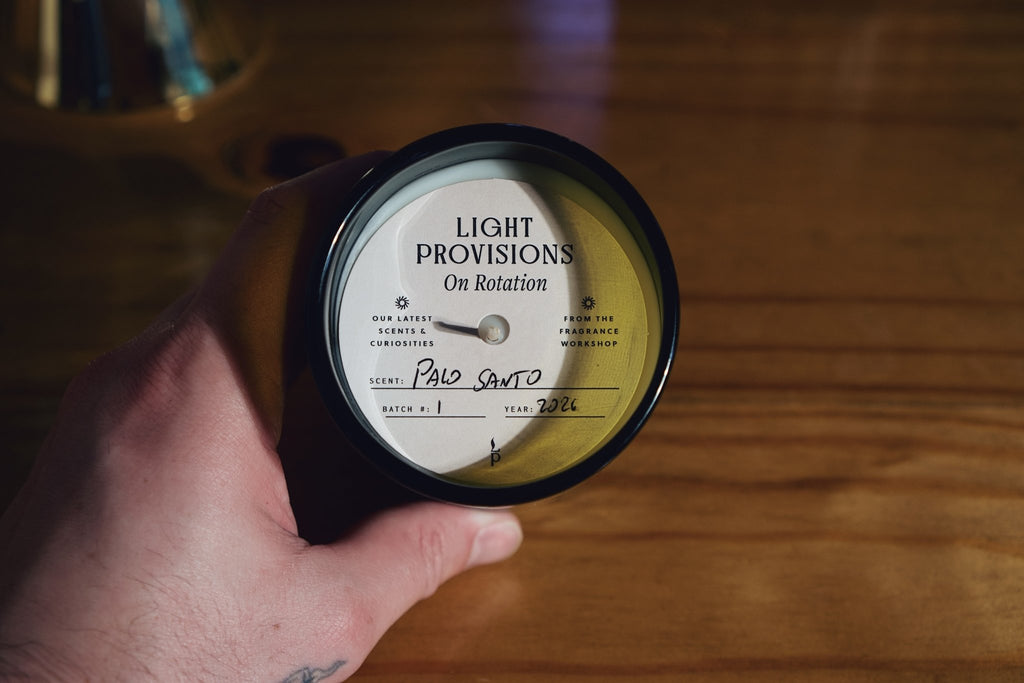 A hand holds a antique green recycled glass vessel with a white label reading 'LIGHT PROVISIONS On Rotation' against a warm wooden background.