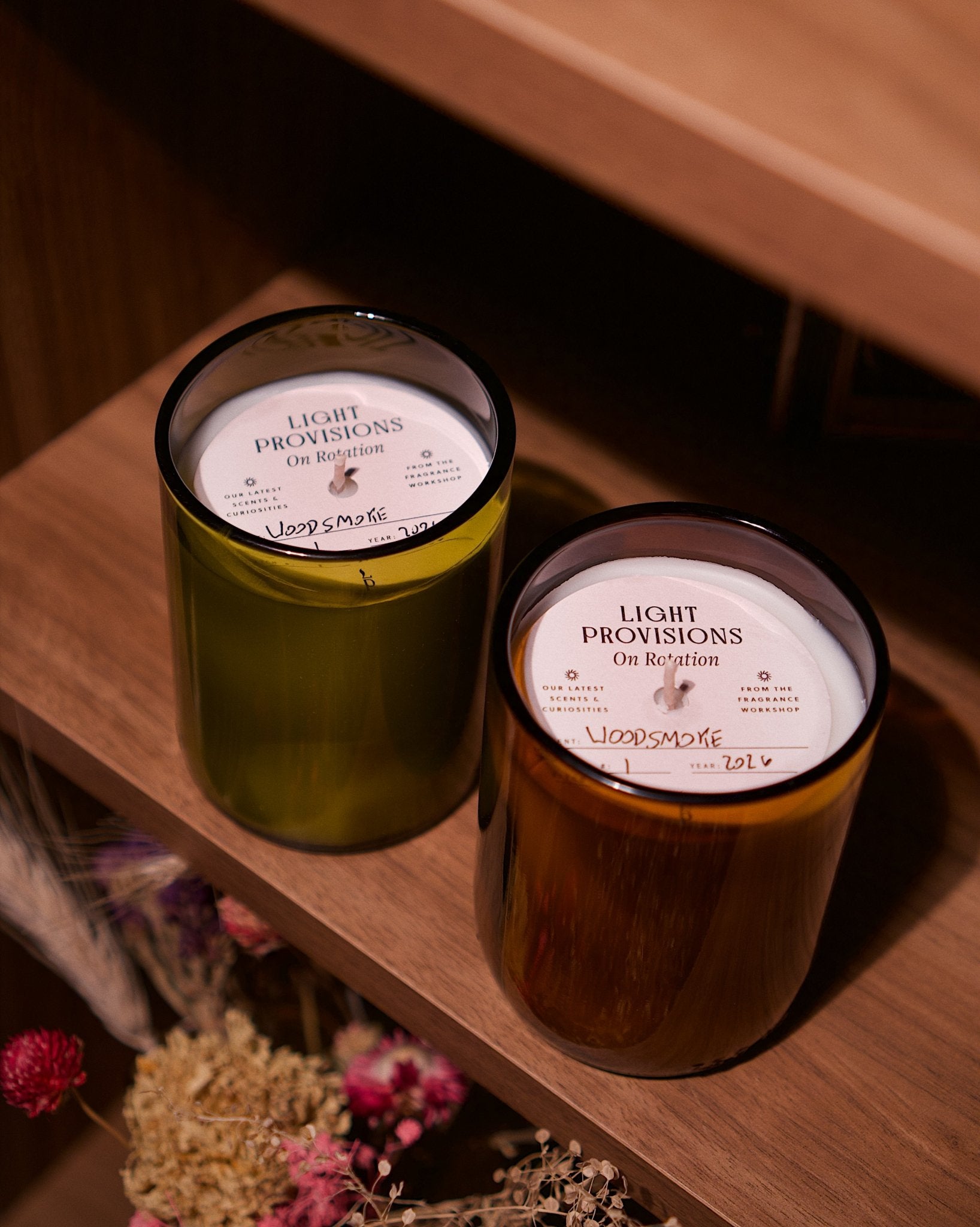 Two glass jar candles with white labels reading 'LIGHT PROVISIONS On Rotation' sit on a wooden shelf. One candle has a yellow-green vessel and the other has an amber-brown recycled vessel, with dried flowers visible in the lower left corner.