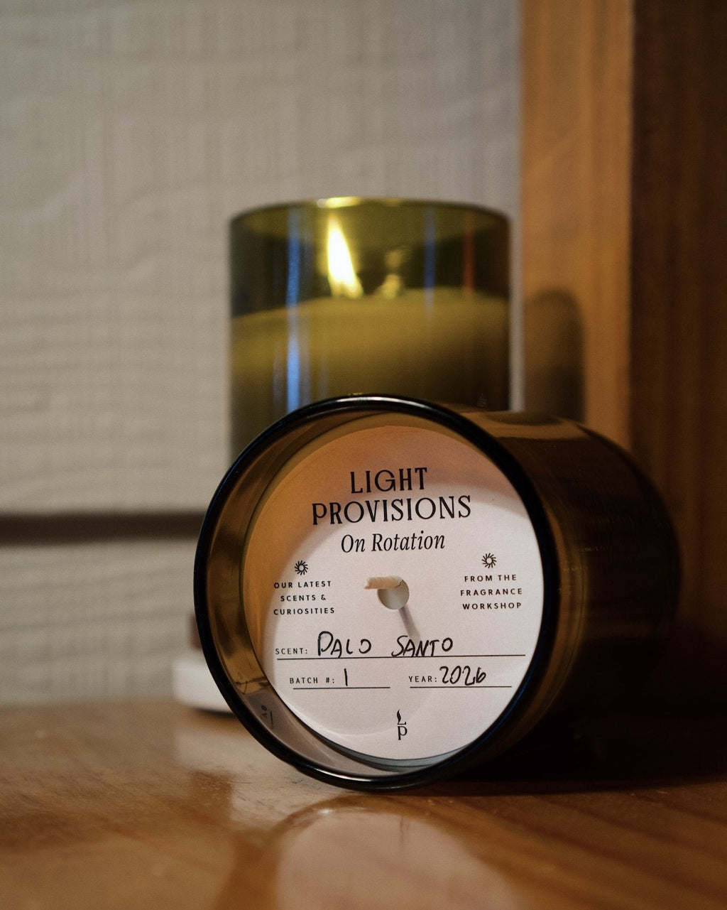 A dark amber glass jar candle lies on its side on a wooden surface, showing the front label that reads 'LIGHT PROVISIONS On Rotation' and 'Palo Santo'. A second candle stands upright in the background in a matching antique green recycled glass container.