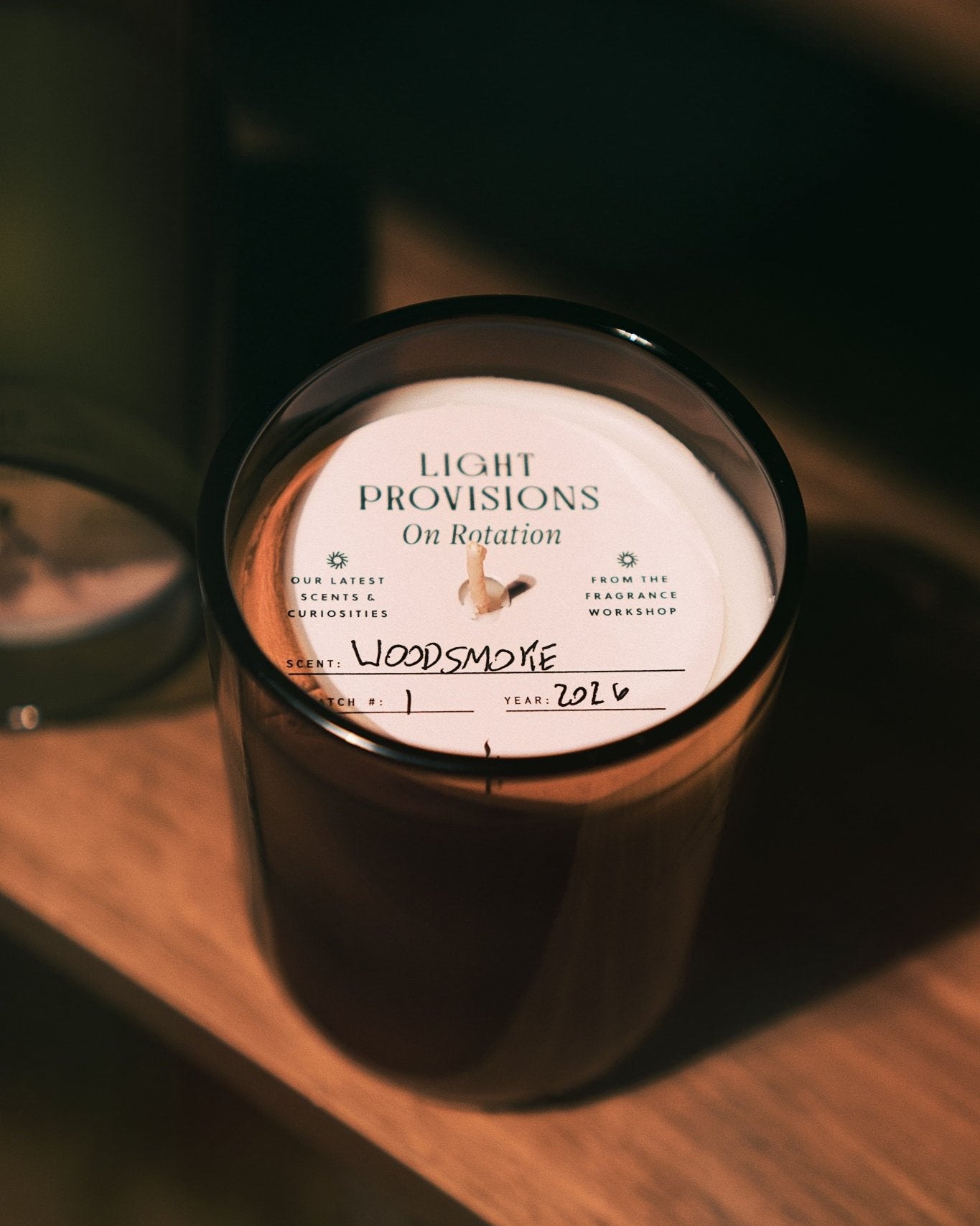 A dark amber glass jar candle on a wooden surface, featuring a white circular label that reads 'LIGHT PROVISIONS On Rotation' with handwritten 'Woodsmoke' text below.