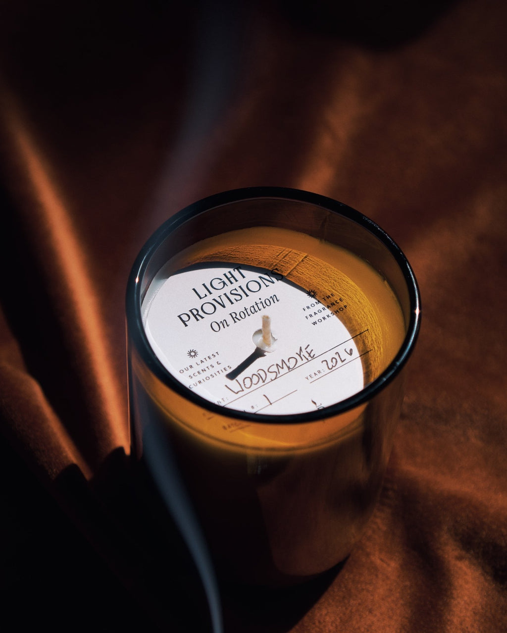 A glass jar candle with amber-colored wax and a white circular label reading 'LIGHT PROVISION On Rotation' and 'WOODSMOKE' photographed on draped brown fabric in warm, low light.