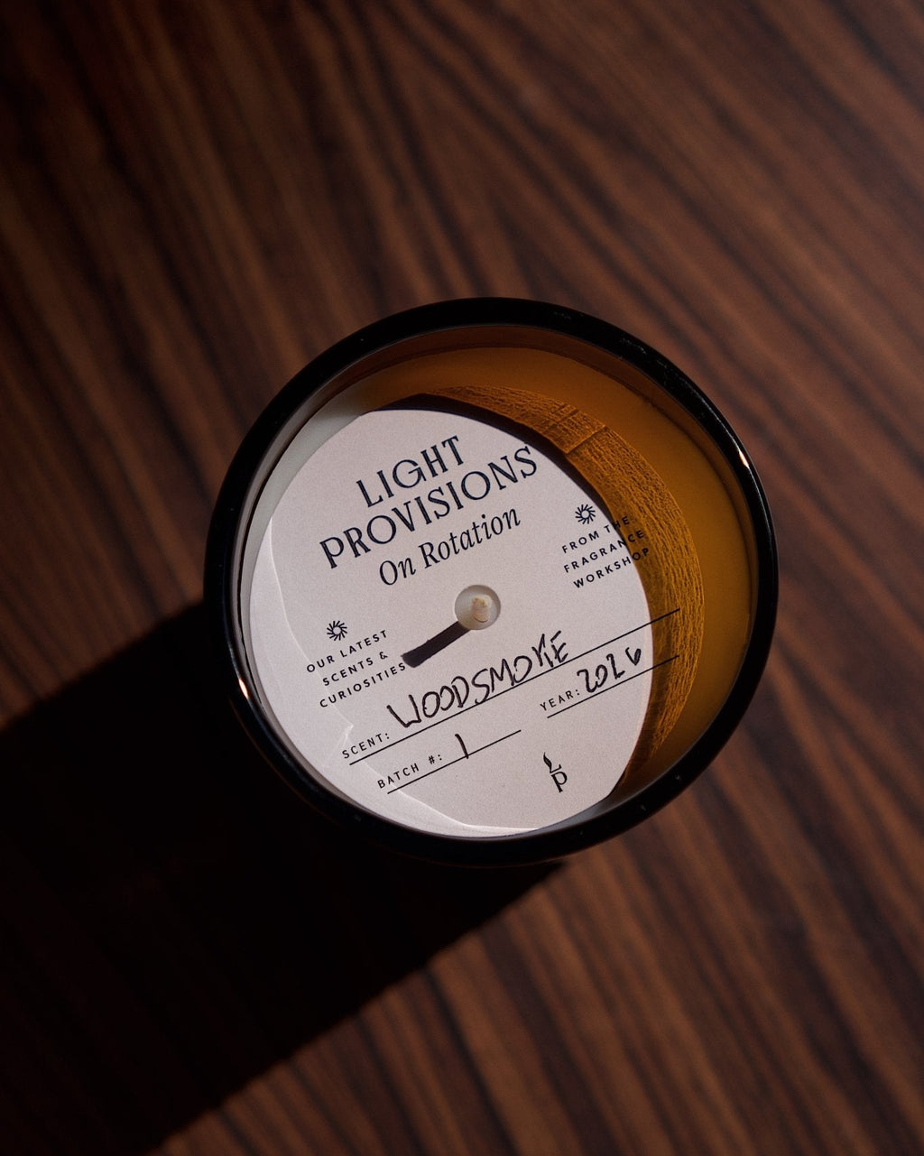 A single candle in a dark recycled glass glass container sits on a dark wood surface. The candle label reads 'LIGHT PROVISIONS On Rotation' and 'WOODSMOKE', with handwritten text 