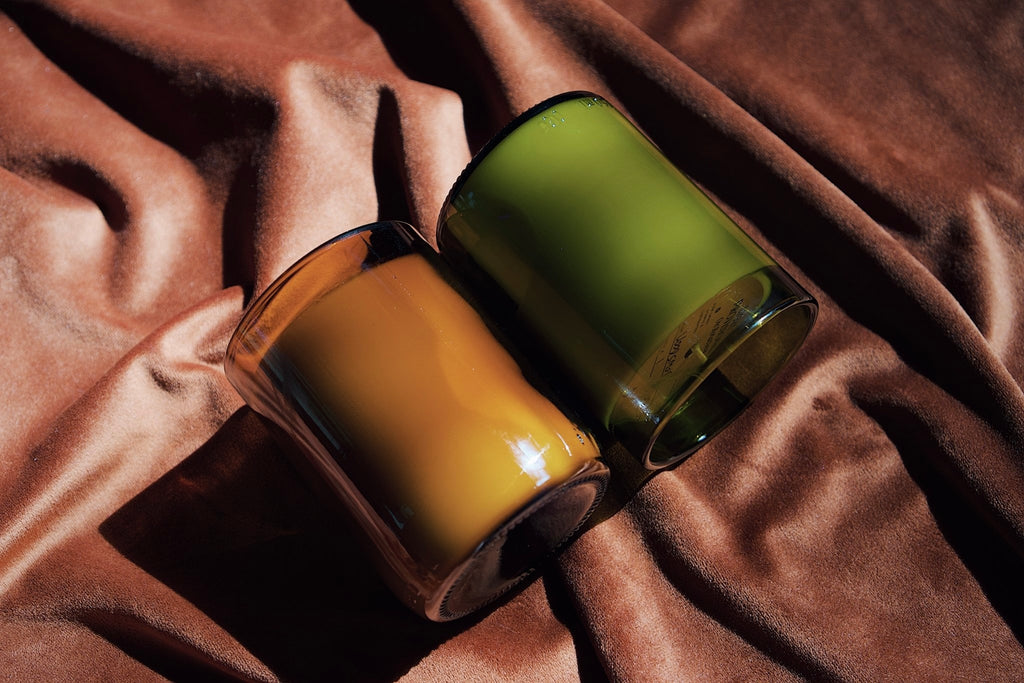 Two recycled wine bottle glass jar candles with amber and olive green colors, resting on draped brown fabric.