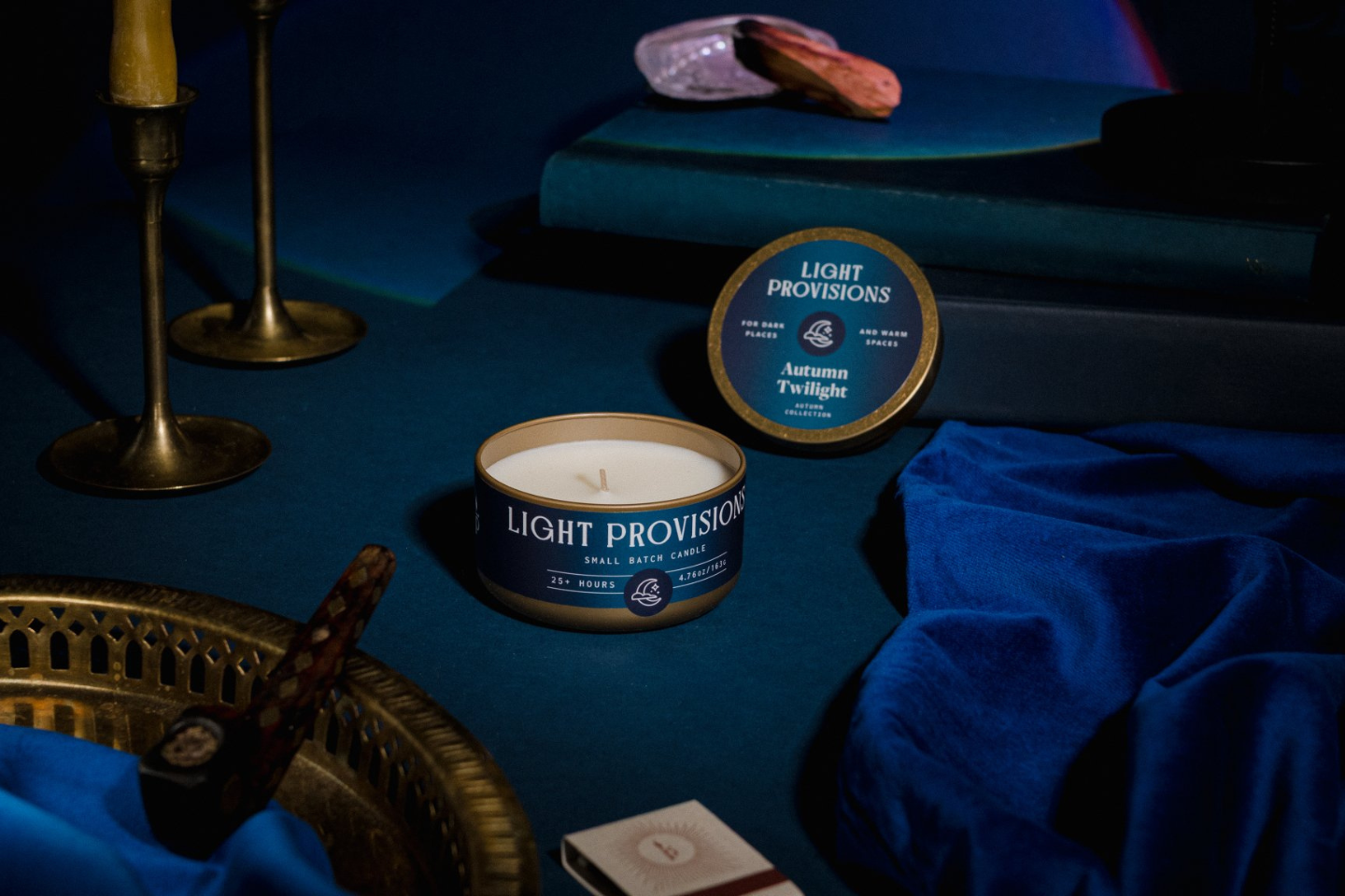 A tin candle labeled Light Provision Autumn Twilight sits on a dark blue velvet surface with its lid beside it. The scene includes brass candlesticks, draped blue fabric, a tray with a cigar, and stacked books in the background, creating a moody styled product presentation.