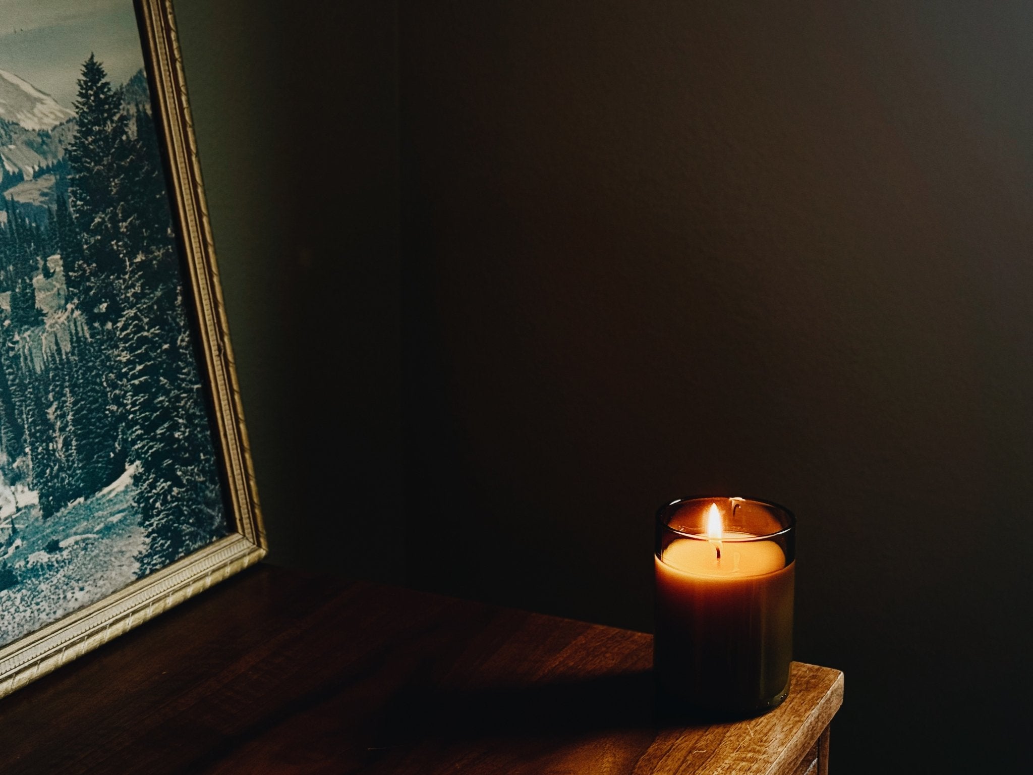 A lit candle in a amber recycled glass sits on a wooden table in a dimly lit room, next to a framed landscape print.