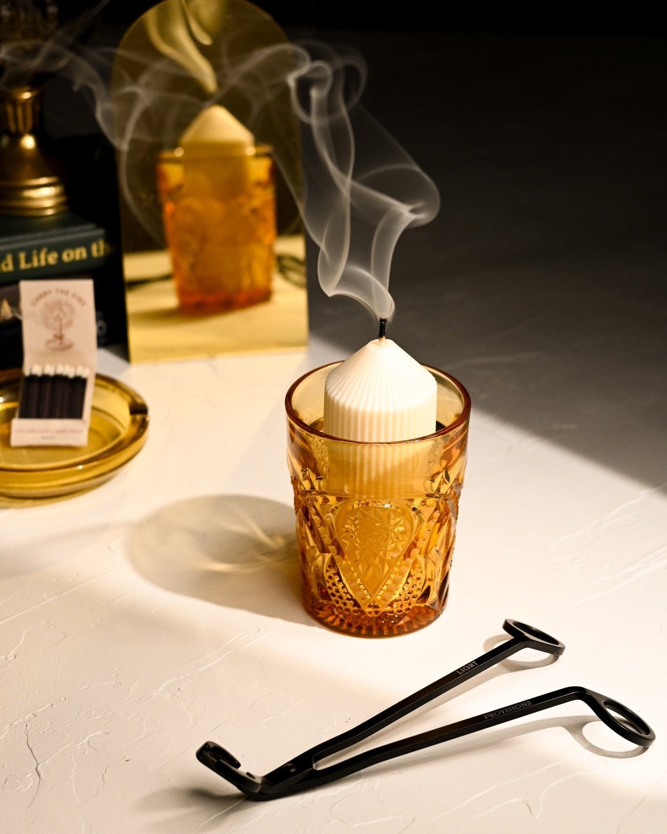 Refill candle in a vintage glass cup with wick trimmer on a table, with a blurred background of bottles and a mirror.