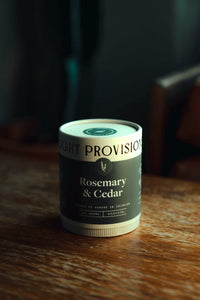 Cylindrical “Light Provisions” Rosemary & Cedar candle tube on a wood table, front label readable moody window light.