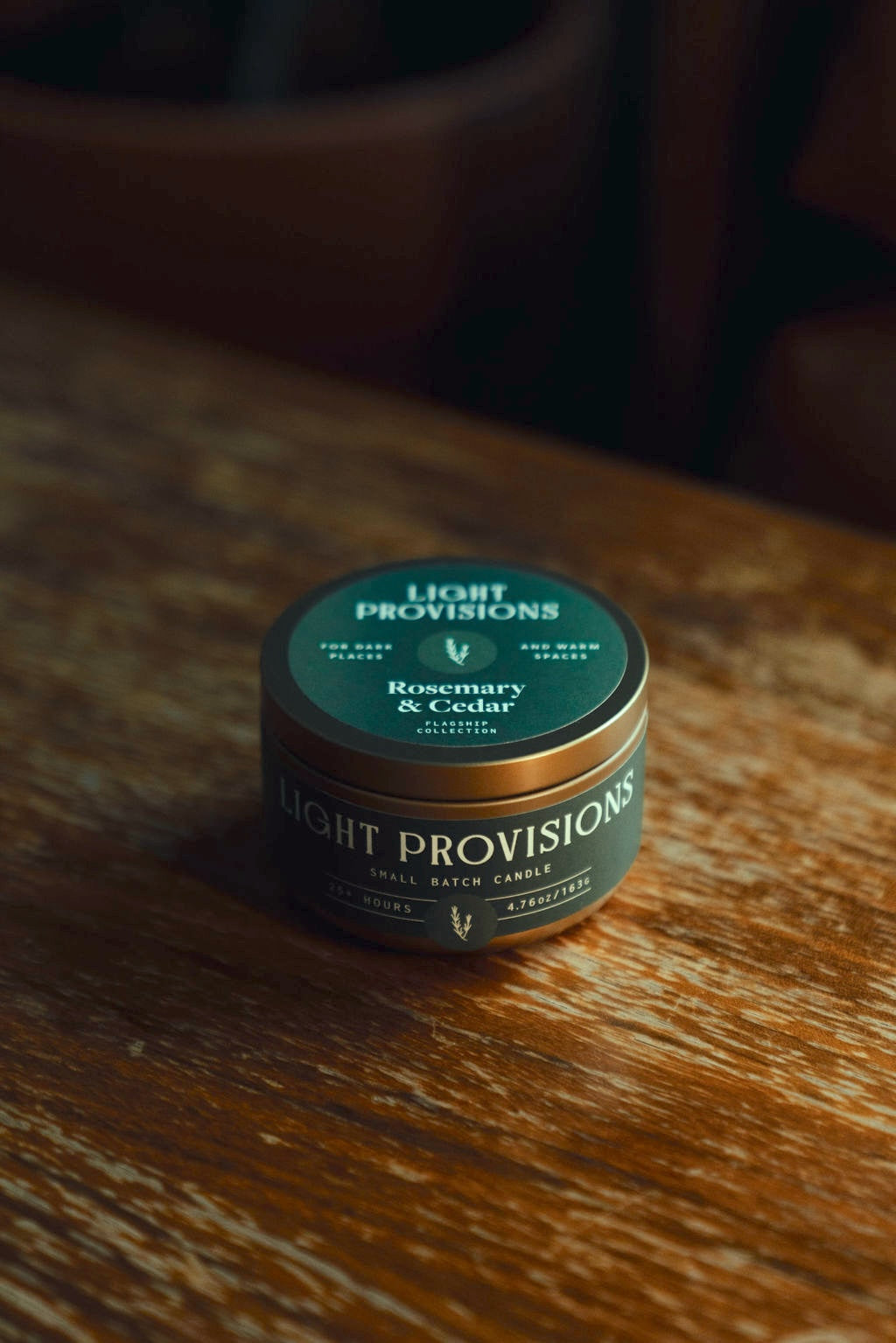 Closed Rosemary & Cedar travel tin on the table; deep-green label, gold lid, “Small Batch Candle · 25+ Hours” visible.