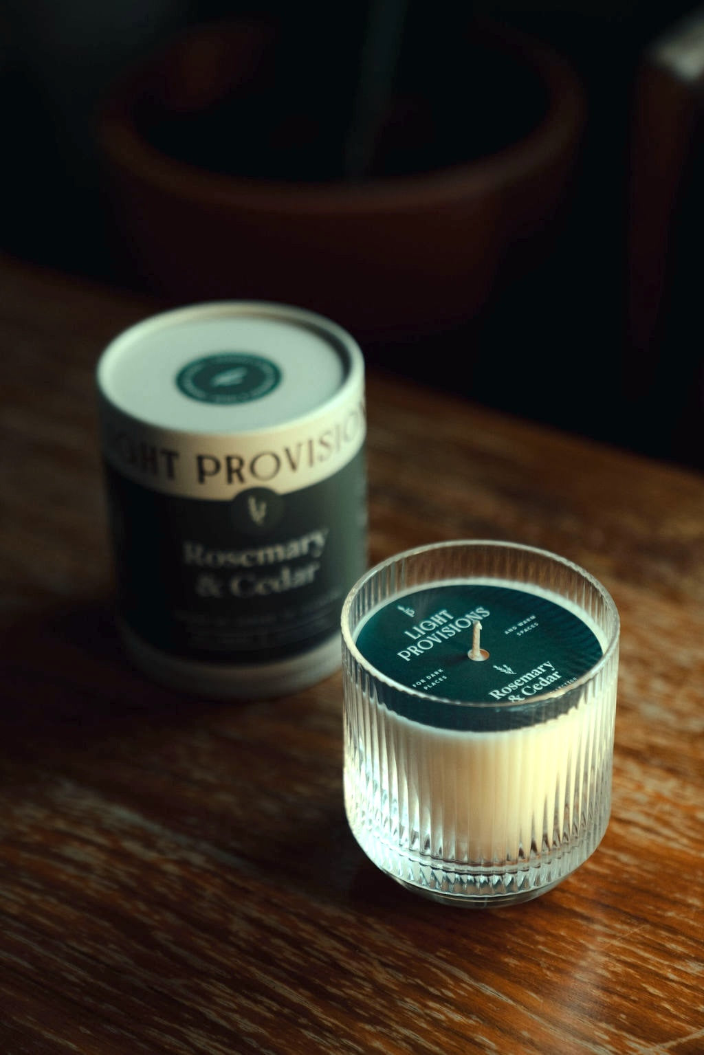 Fluted glass tumbler candle in foreground with tube blurred behind;dust cover and centered wick visible through the glass.