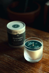 Fluted glass tumbler candle in foreground with tube blurred behind;dust cover and centered wick visible through the glass.