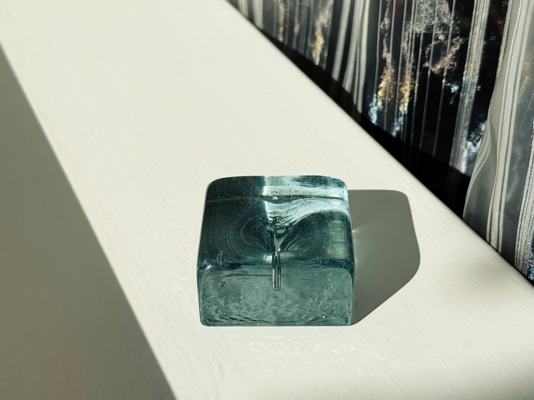 A small translucent teal-blue square glass block sculpture with a central vertical crack, resting on a white surface with a strong shadow.