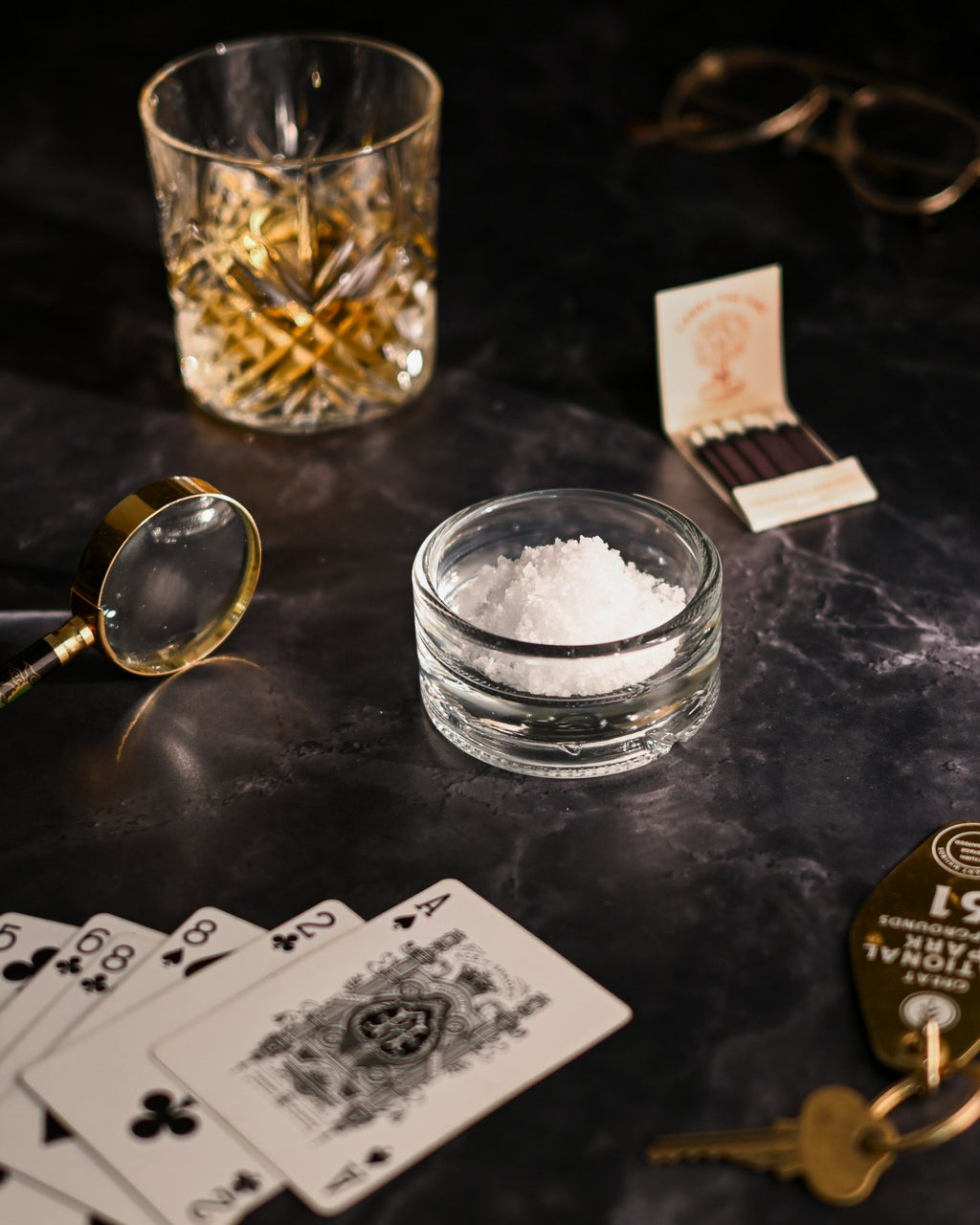 Clear reclaimed bottle glass dish used as a small salt dish. whiskey glass, matches, magnifying glass, and playing cards in background