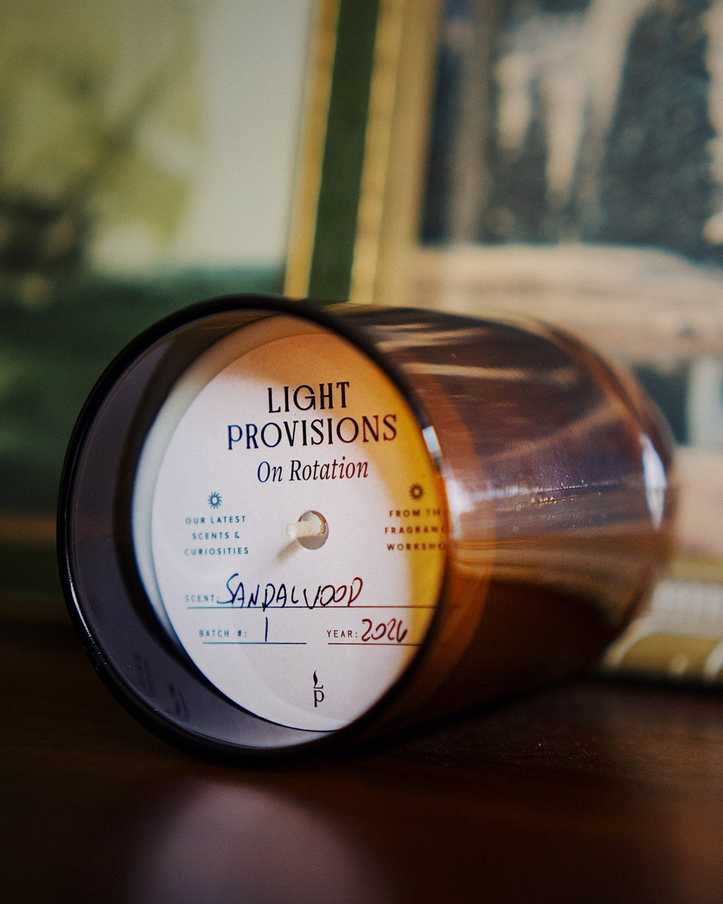 A close-up of a lit soy wax candle in a dark amber glass jar, lying on its side on a dark wooden surface. The white circular label reads 'LIGHT PROVISIONS On Rotation' and is handwritten with 'Sandalwood' in blue ink.