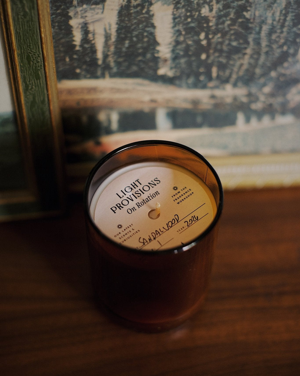 A soy wax candle in a dark amber glass jar sits on a wooden table in front of a framed winter landscape print. 
