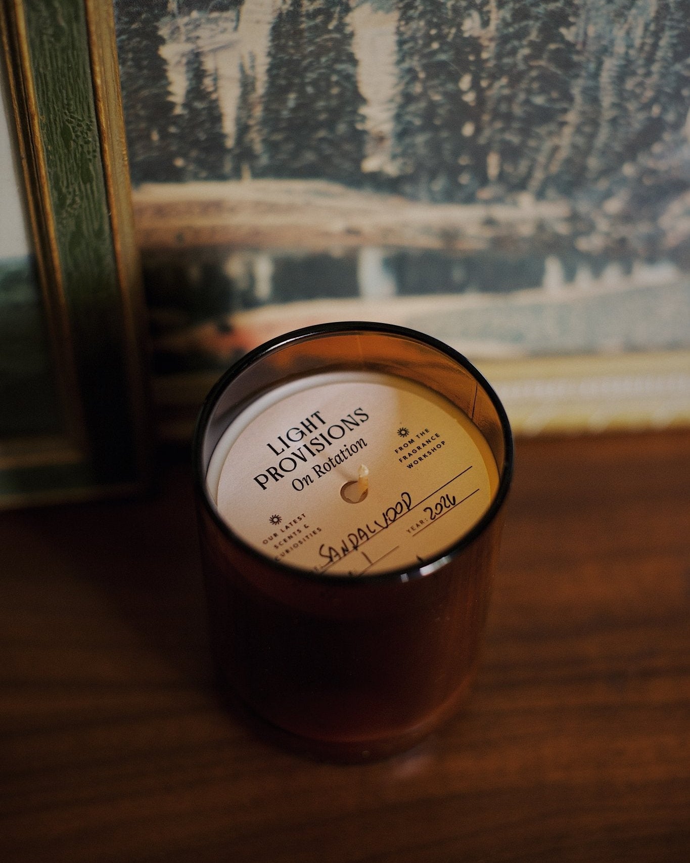 A soy wax candle in a dark amber glass jar sits on a wooden table in front of a framed winter landscape print. 