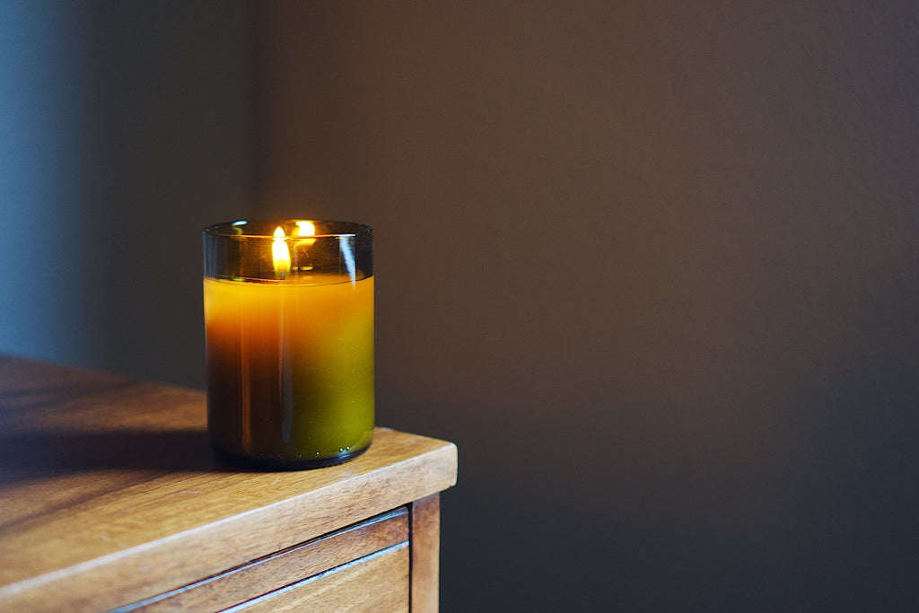 A lit candle in a antique green recycled glass jar sits on a wooden surface against a dark brown wall.