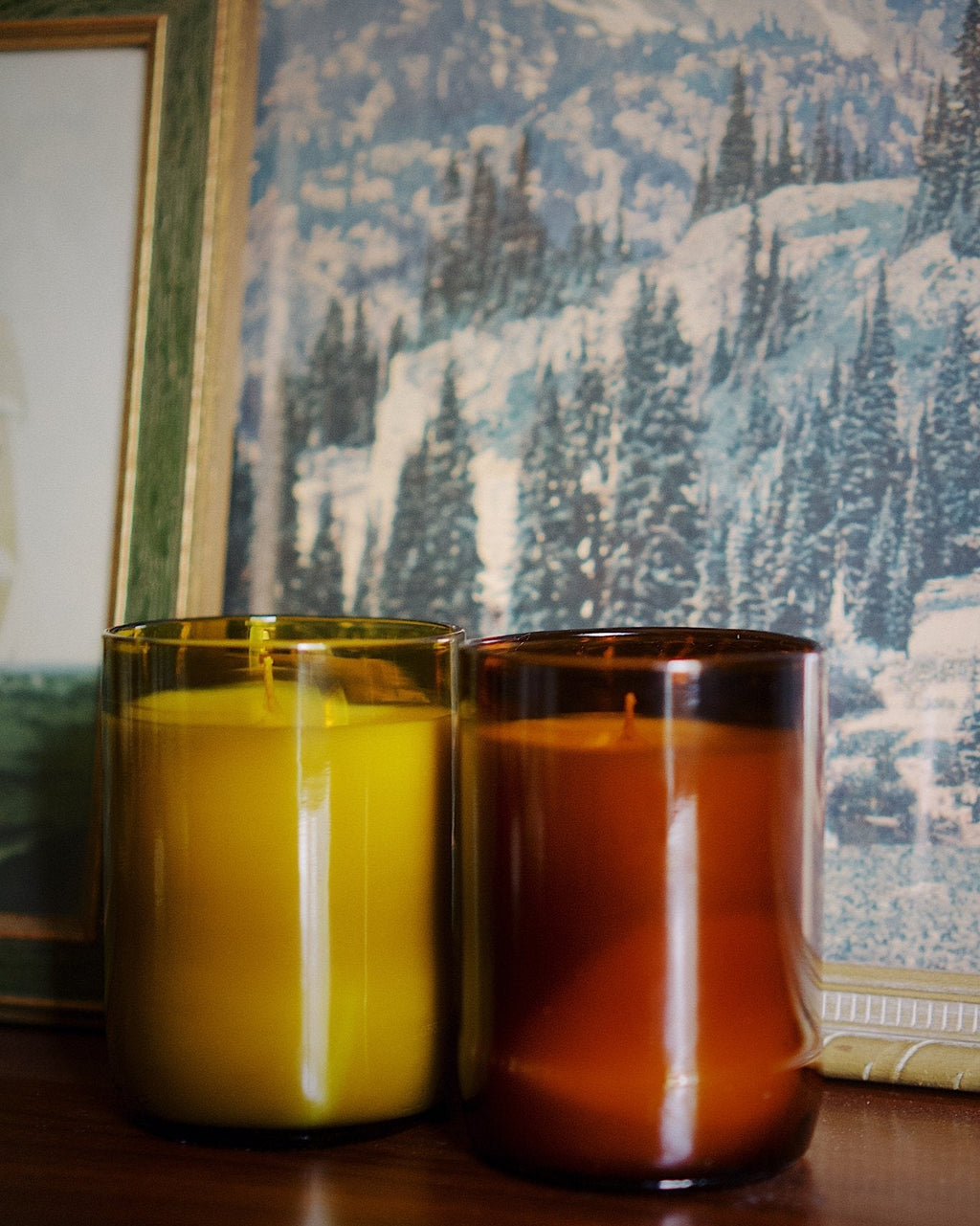 Two amber glass jar candles, one antique green and one amber, sitting on a dark wooden surface in front of a scenic winter mountain wallpaper and a framed picture.