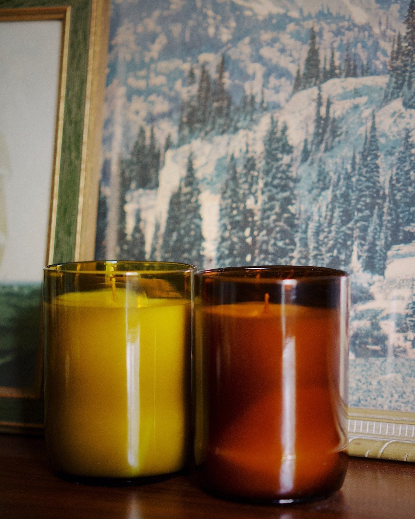 Two amber glass jar candles, one antique green and one amber, sitting on a dark wooden surface in front of a scenic winter mountain wallpaper and a framed picture.