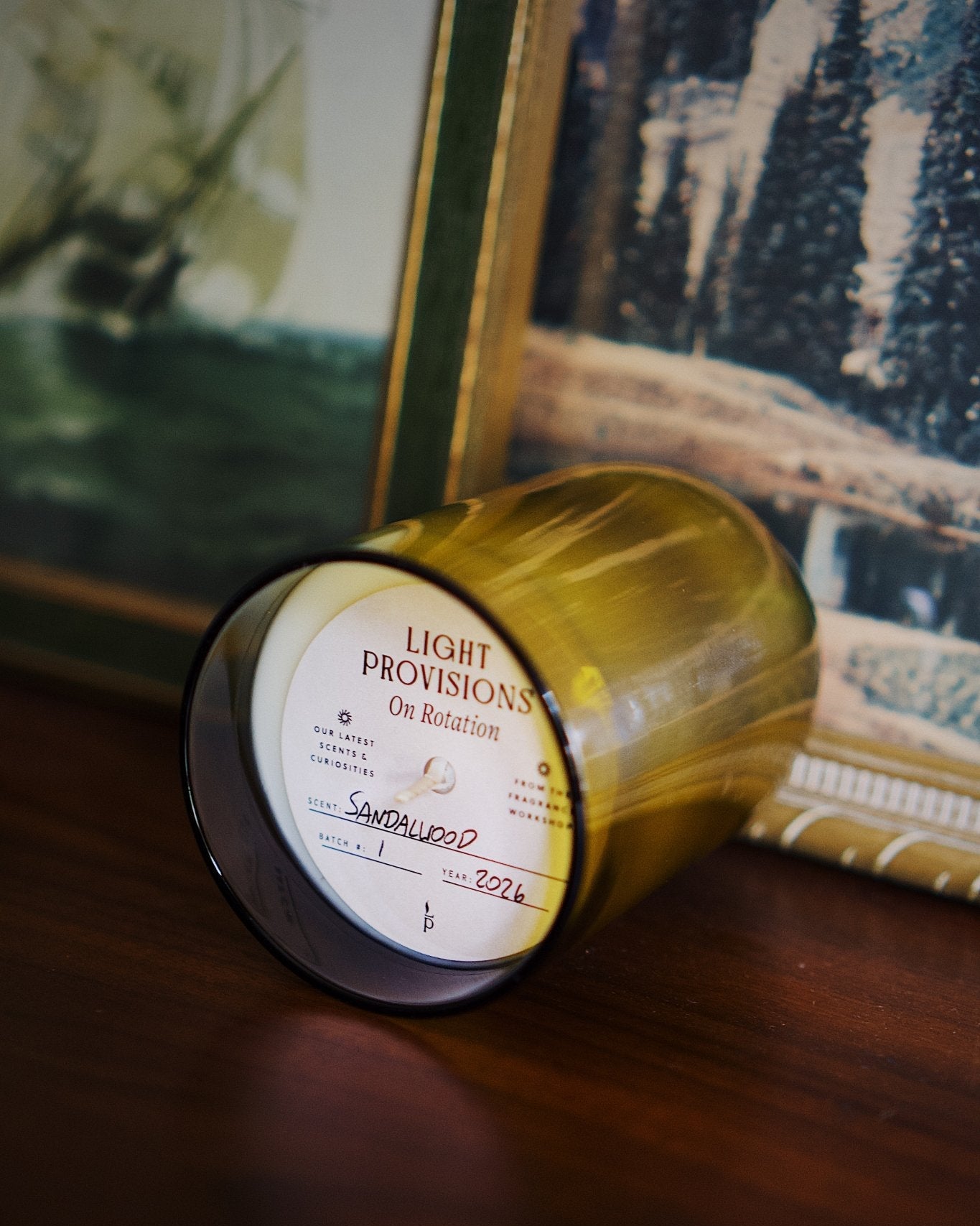 A candle in a translucent antique green recycled glass jar lies on its side on a dark wooden surface, with a white label reading 'LIGHT PROVISIONS and Sandalwood; visible. Framed artwork and a notebook are blurred in the background.