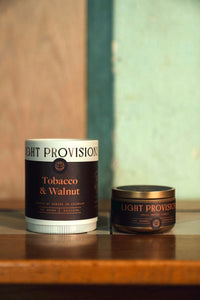 Light Provisions Tobacco & Walnut candle packaging and travel tin displayed together on wooden surface.