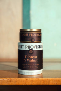 Tobacco & Walnut travel tin candle stacked on top of tumbler packaging, both with matching brown and copper foil labels.