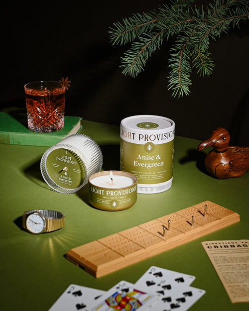 Anise & Evergreen candle with packaging, playing cards, and a drink on green tabletop under pine branches