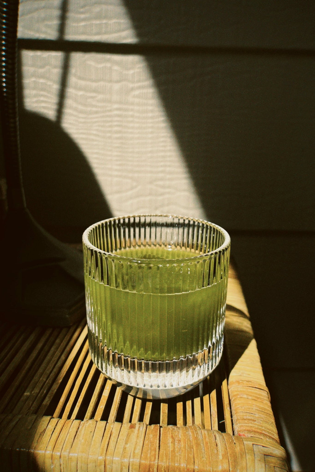 Ribbed glass tumbler holding a pale green matcha latte with ice, resting on a woven chair seat in afternoon sunlight.