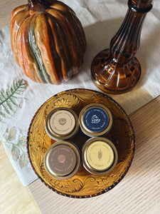 Four sample-sized autumn tin candles with lids on a decorative glass plate, surrounded by a ceramic pumpkin and a glass container on a textured surface.
