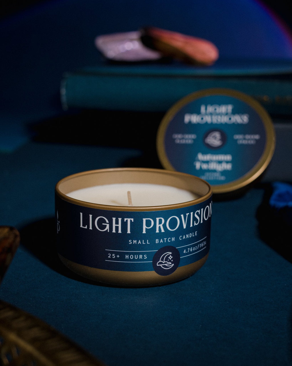 Autumn Twilight candle travel tin open with warm glow on dark blue tabletop with books and velvet