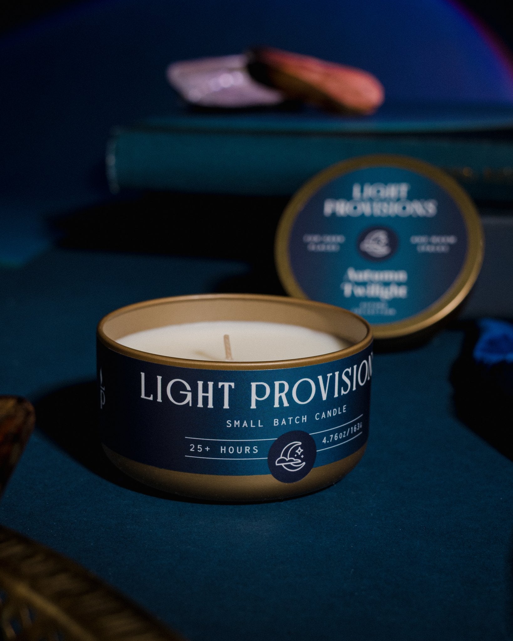 Autumn Twilight candle travel tin open with warm glow on dark blue tabletop with books and velvet