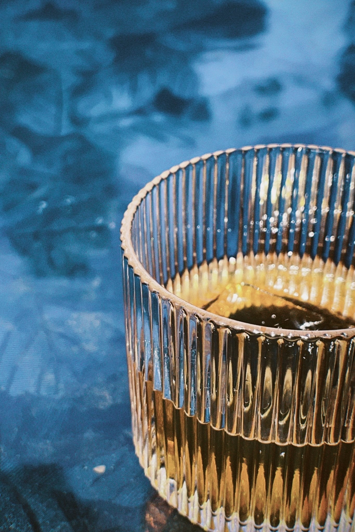 Ribbed tumbler filled with a light golden cocktail, photographed on a blue textured surface in natural light.