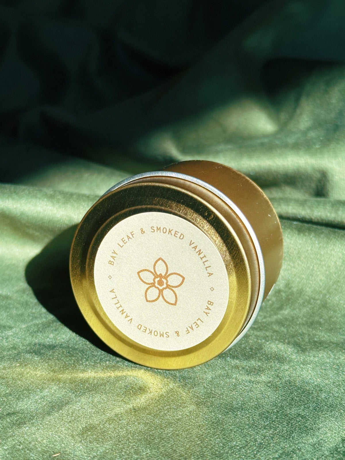 Gold sample tin labeled Bay Leaf and Smoked Vanilla resting on green fabric with direct sunlight.