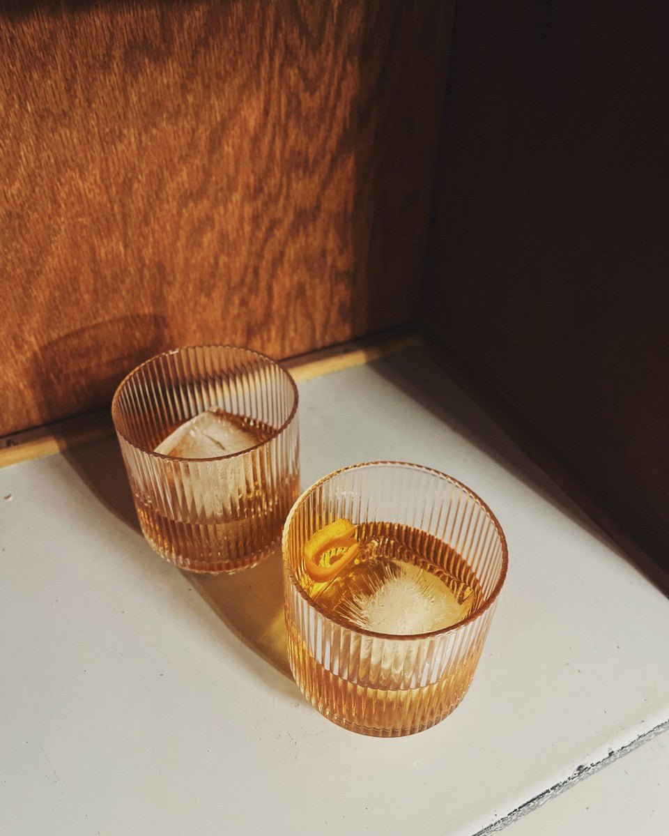 Bespoke glass tumblers with shots of whiskey, photographed in warm sunlight on a white surface beside wood paneling.