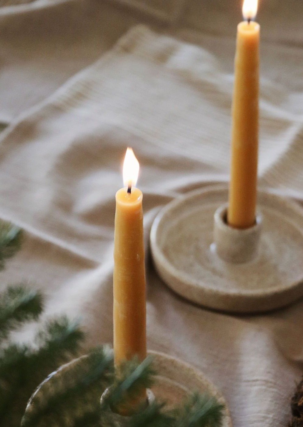 2 beeswax candles burning in ceramic holders on a linen surface with pine needle decor in the background