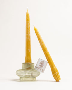 Light Provisions hand-dipped beeswax taper candles, one in a ribbed glass holder and one leaning, natural bloom visible.