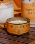 Bitter Orange & Clove tin candle resting on a striped wood tabletop beside the larger glass candle and packaging tube.