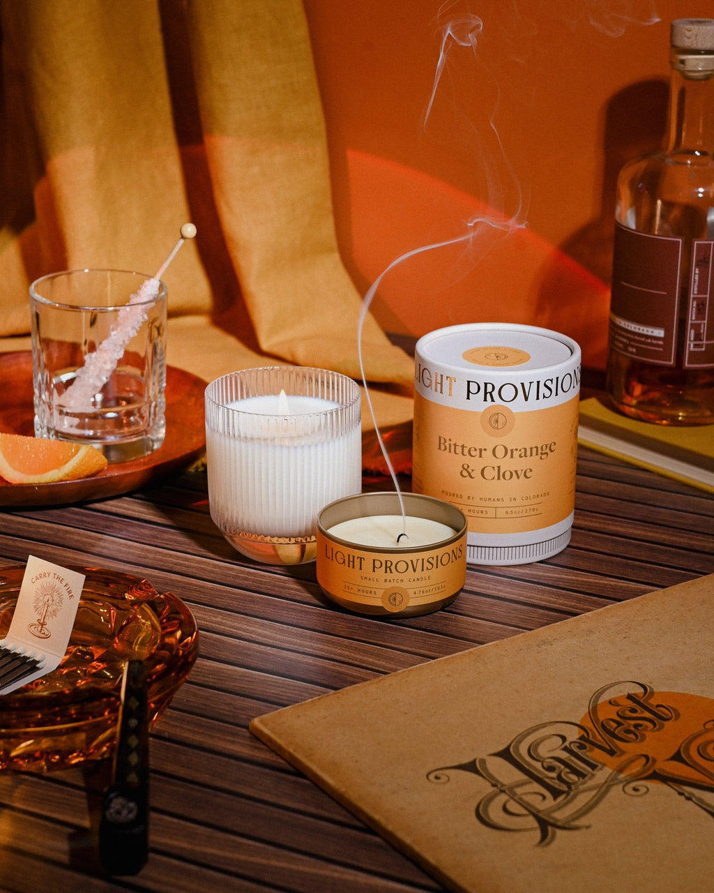 Bitter Orange & Clove candles with whiskey glass and notebook, golden hour tones highlighting wood and textile textures.