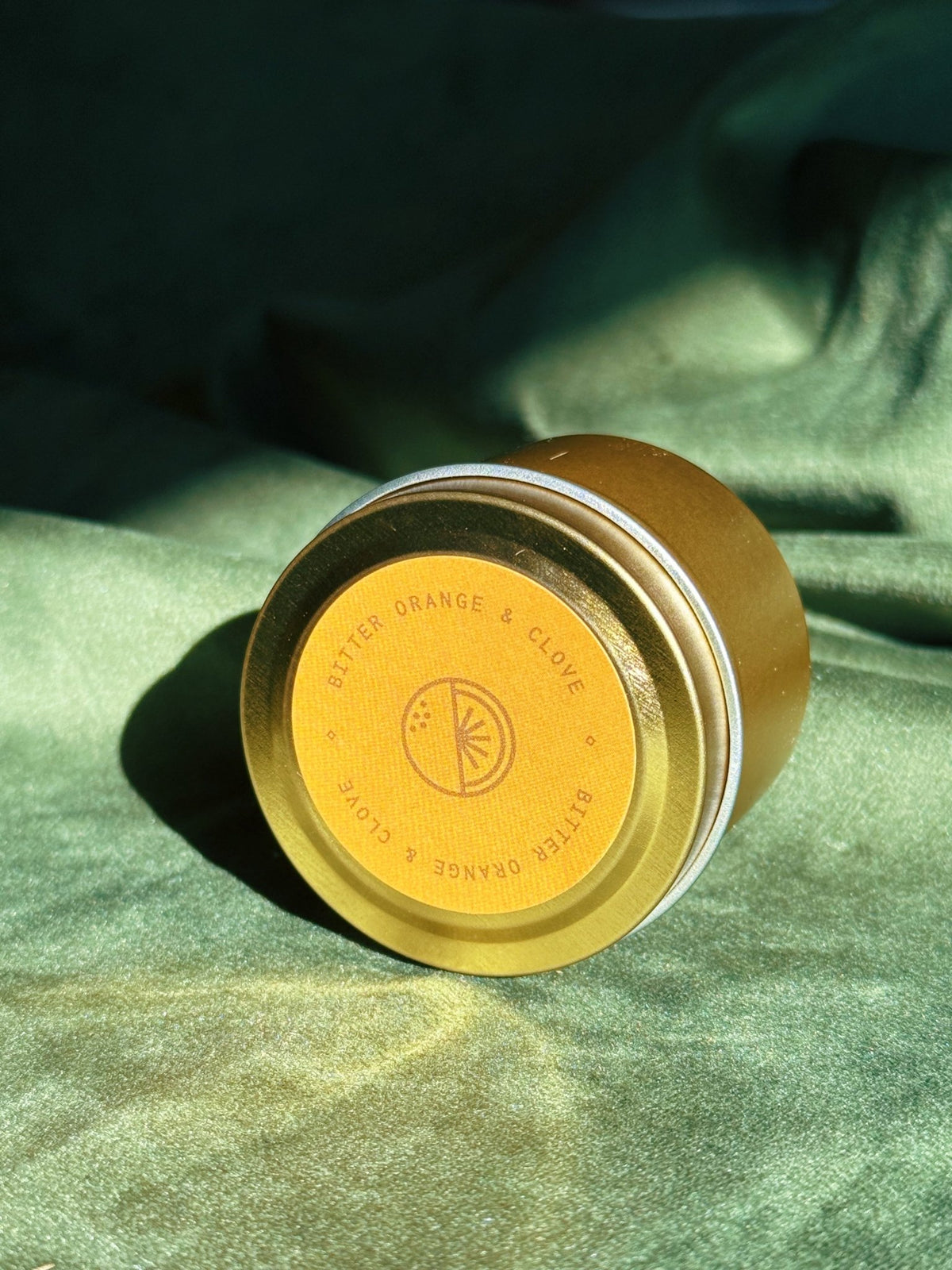 Gold sample tin labeled Bitter Orange and Clove resting on green fabric in sunlight.