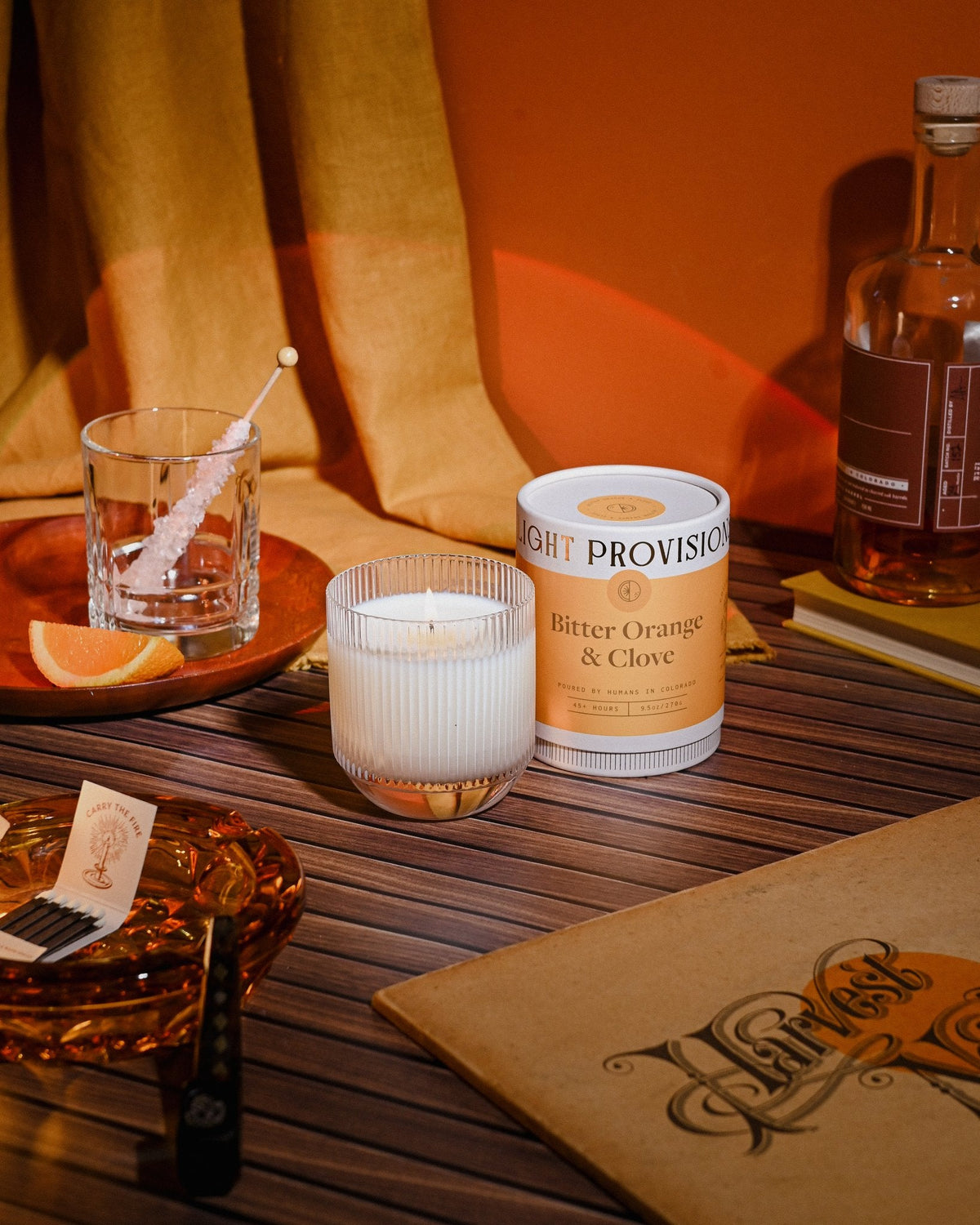 Bitter Orange & Clove tumbler candle burning, orange glow reflecting on amber drinkware and fabric backdrop.