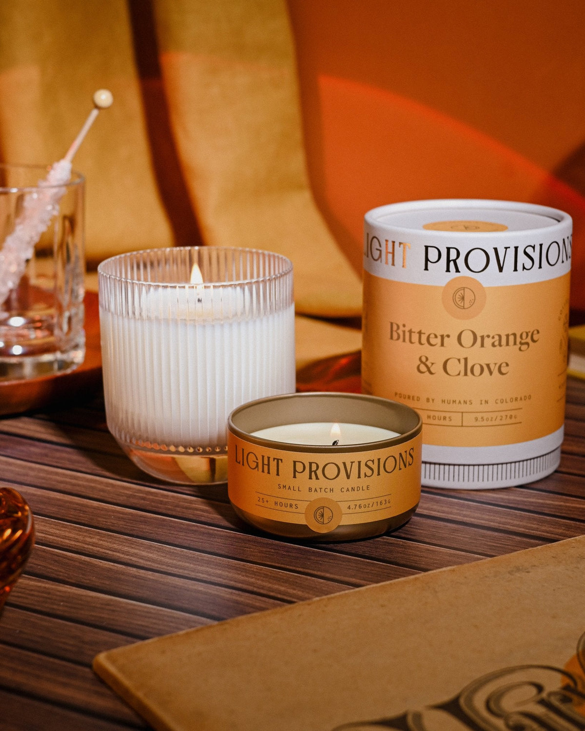 Candle beside a ribbed glass tumbler and cocktail, warm golden light casting soft shadows on wood grain.