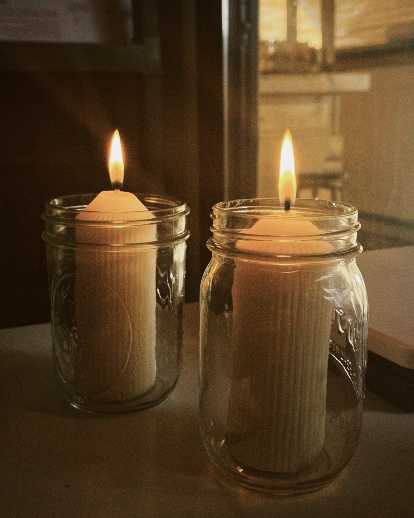 Two lit refill candles in glass mason jars on a reflective surface with a blurred background