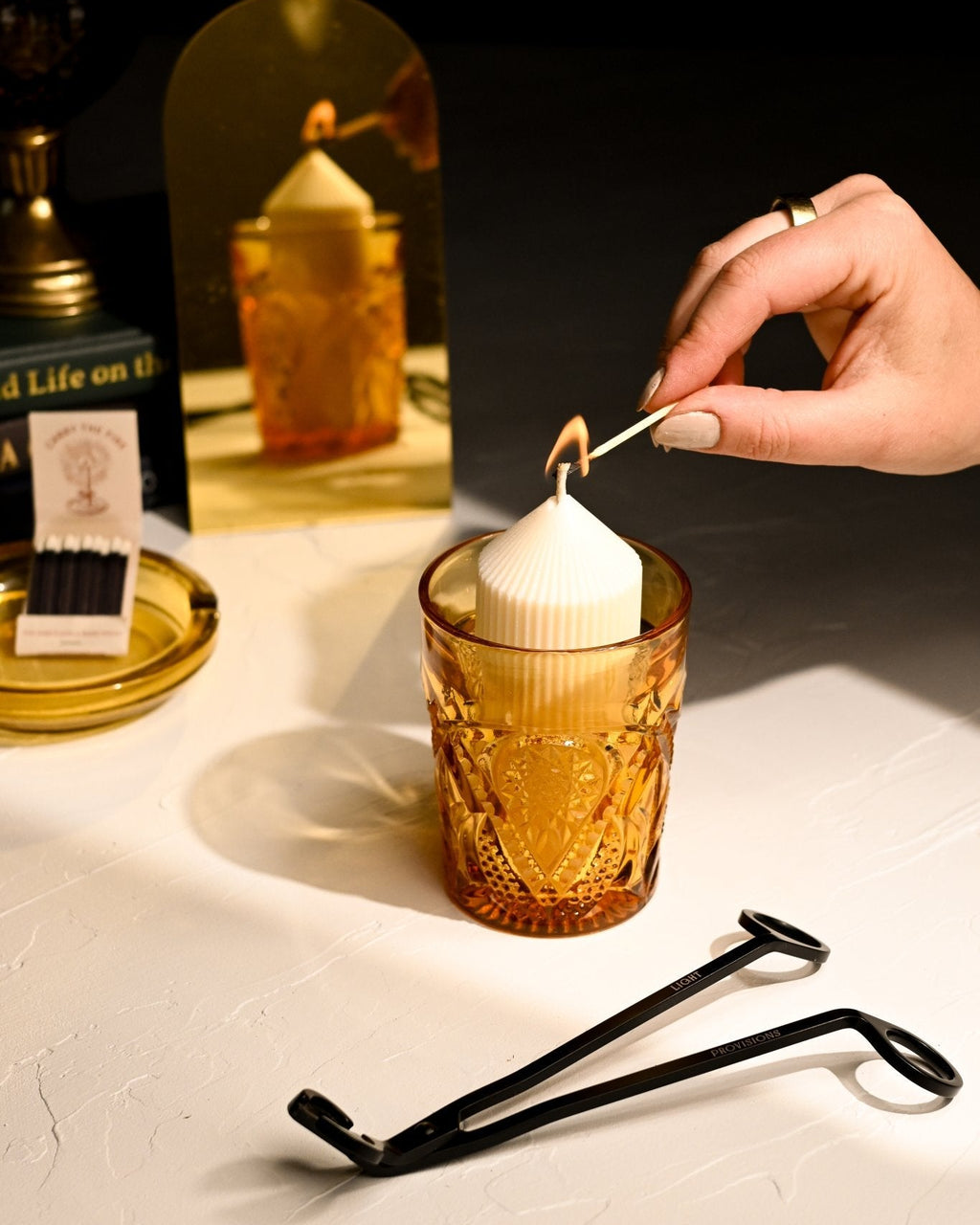 Hand lighting a candle with a match on a table with a glass and tongs.