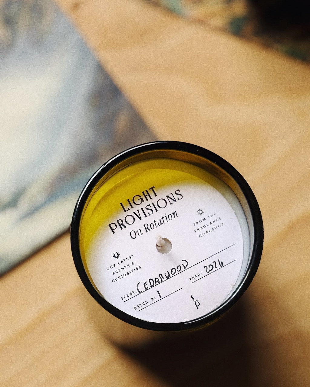 Top-down view of a candle in a black glass jar with yellow wax and a white label reading 'Light Provisions On Rotation' on a wooden surface.