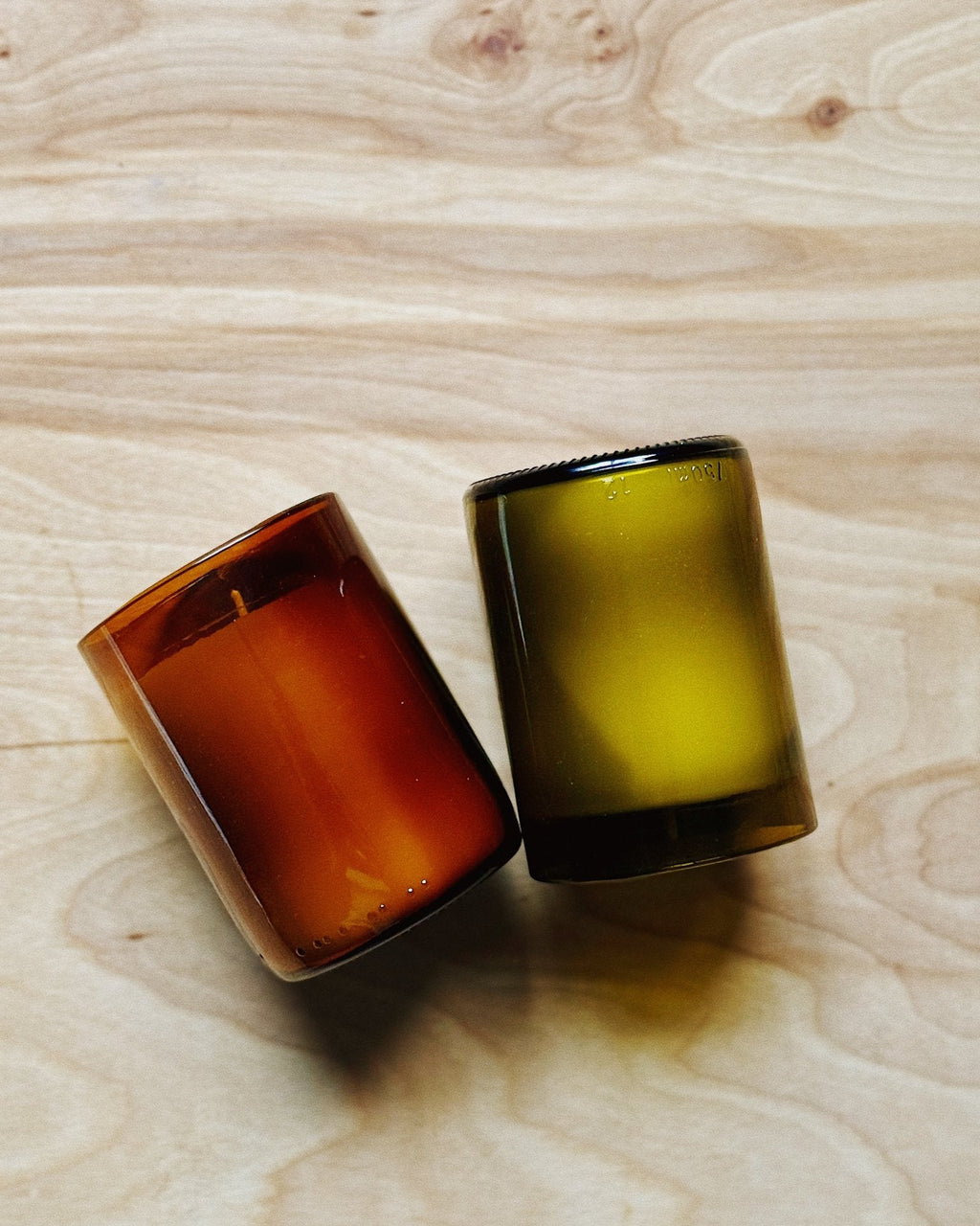 Two recycled glass candle vessels on a light wood grain surface, one translucent reddish-brown and the other olive green.